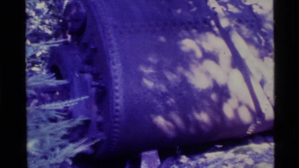 1977 Alaska 8mm frame showing a weathered, riveted cylindrical metal surface beside evergreen growth.