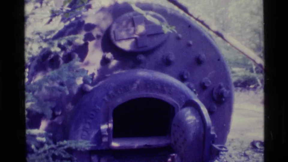 1977 Alaska 8mm frame of a riveted boiler-like front, circular hatch detail, and black arched opening in a wooded setting.