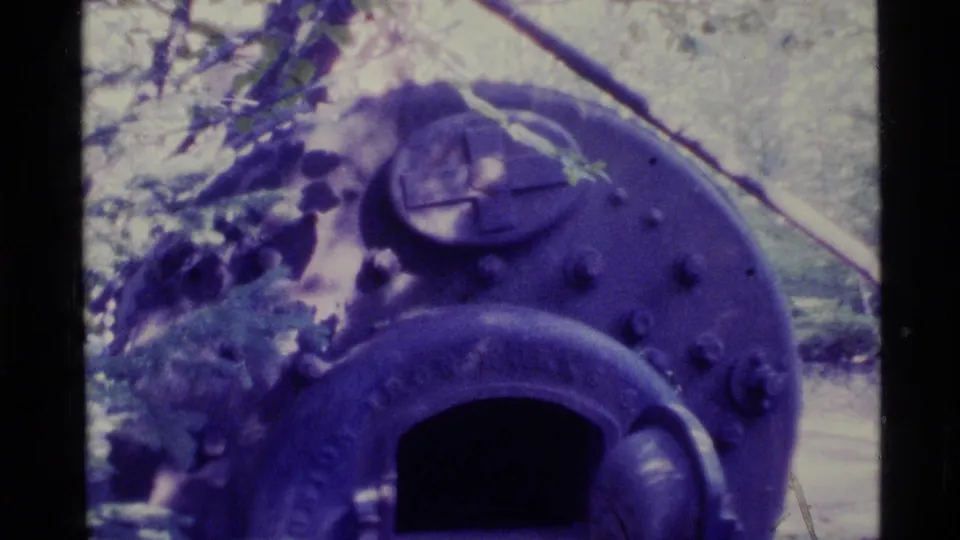 1977 Alaska 8mm frame showing a dark rounded metal stove or furnace near trees, with soft focus and purple color cast.