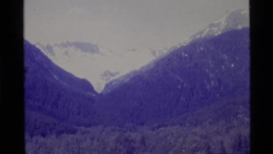 1977 Alaska 8mm frame with snow-bright mountain ridges above dark tree-covered slopes and visible film gate edges.