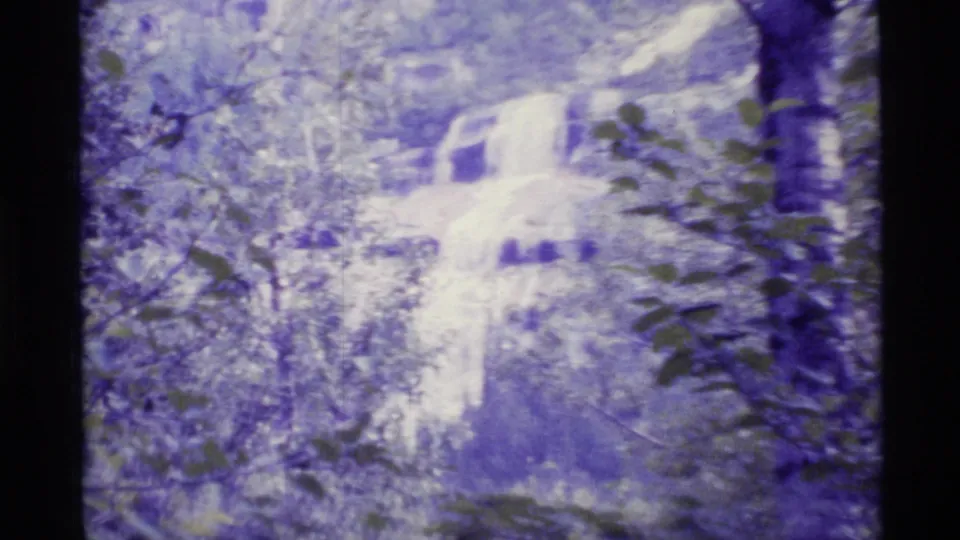 1977 Alaska 8mm frame with a pale cascading waterfall seen through wooded foliage and soft faded color.