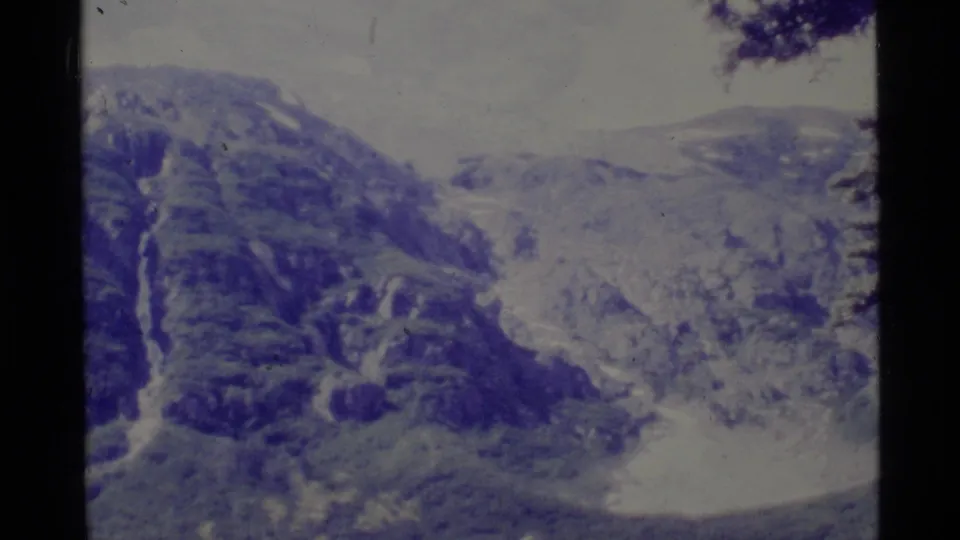 1977 Alaska 8mm wide view of a rugged mountain backdrop above dense forest and a pale stream or trail area.