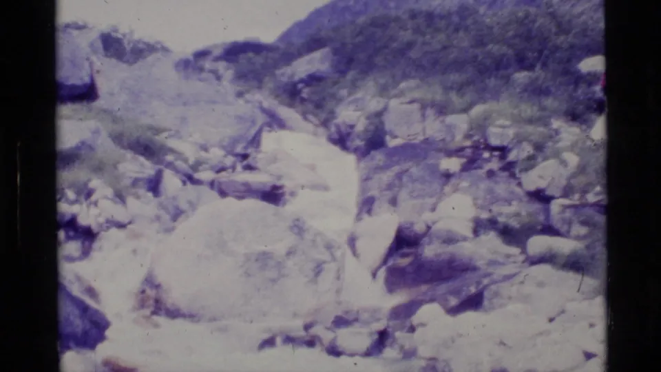1977 Alaska outdoor frame showing a rocky watercourse and hillside vegetation in soft 8mm exposure.