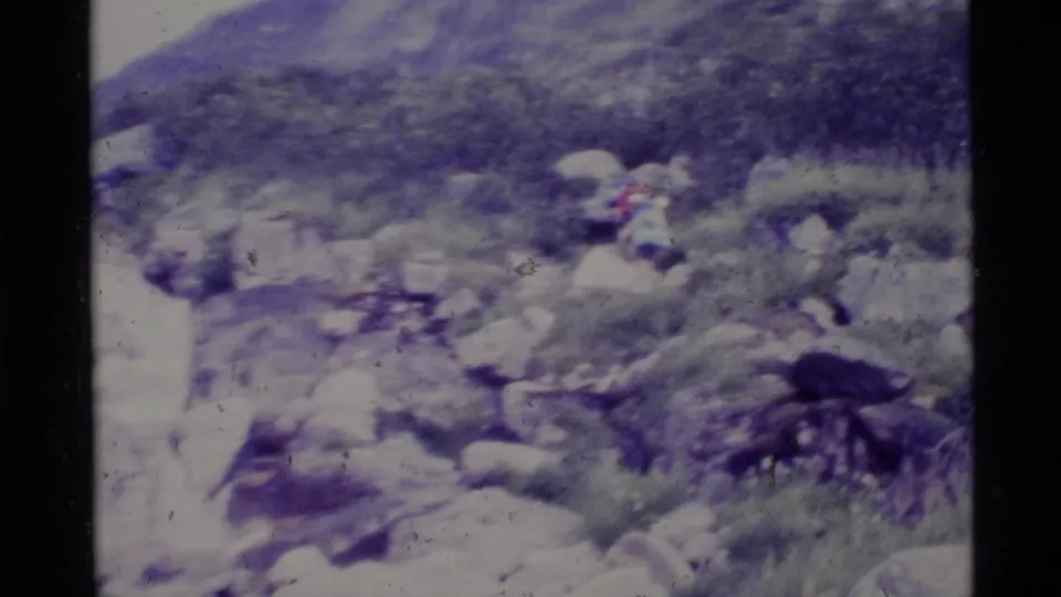 Alaska 1977 8mm frame of a rocky stream area, hillside brush, and a distant figure too small to identify.
