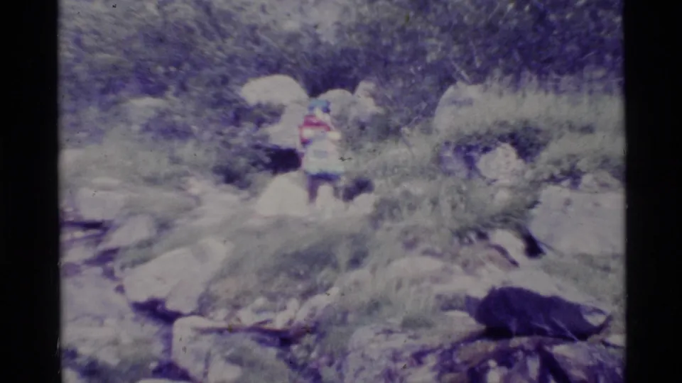 1977 Alaska home-movie frame showing a distant person in bright clothing among rocks and vegetation on rugged terrain.