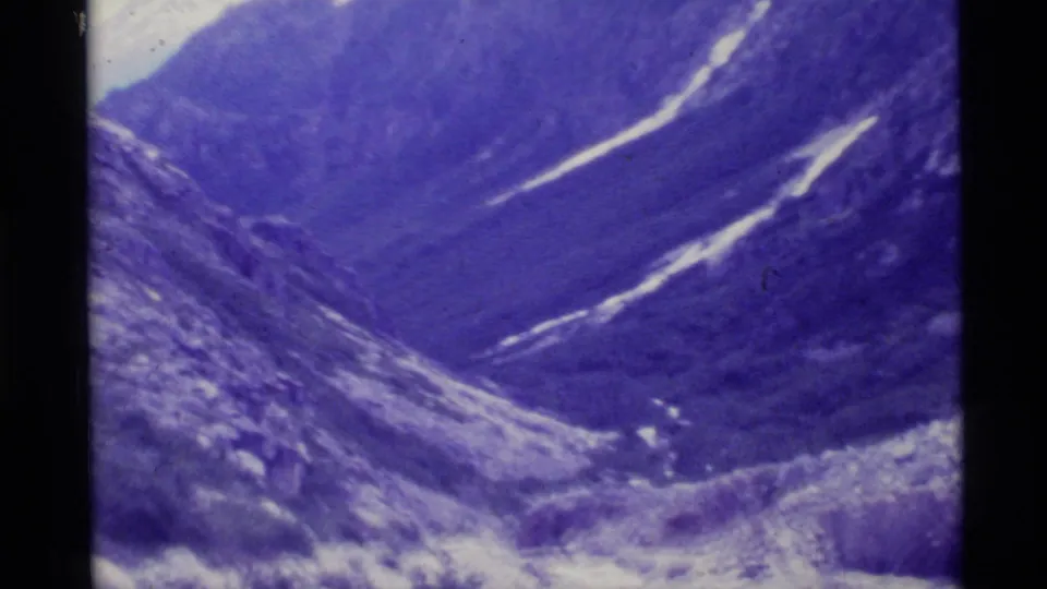 1977 Alaska home-movie frame of a wide mountain valley, seen from a high rocky slope with snow streaks on the opposite side.