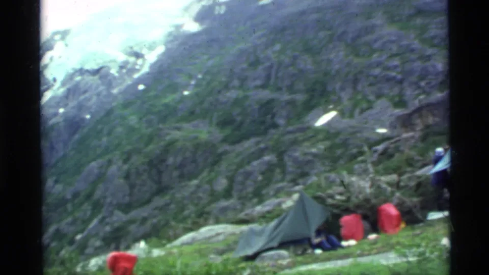 1977 Alaska camping scene with a green tent, red packs, and distant people on a grassy mountainside beneath rocky peaks.