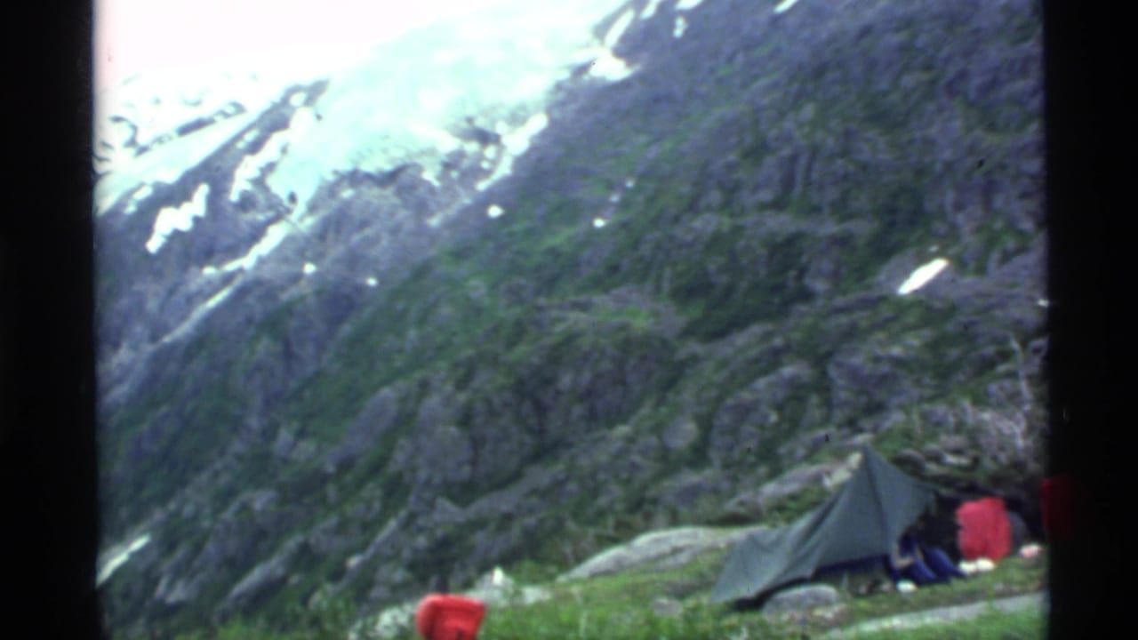 Mountain campsite with a dark tarp shelter, red gear, and rocky snow-streaked slopes behind it.