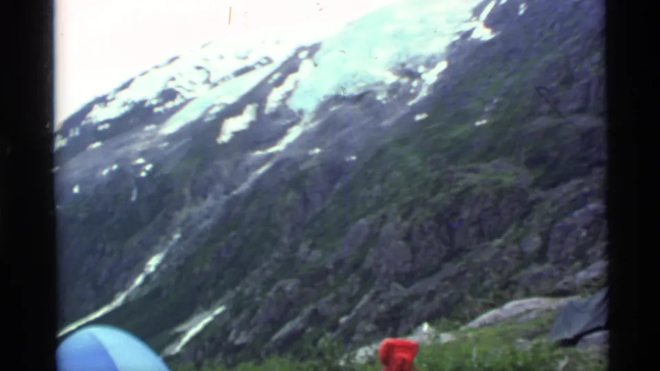 1977 Alaska frame with steep rocky slopes, lingering snow patches, red camping gear, and part of a tent or shelter at the right edge.