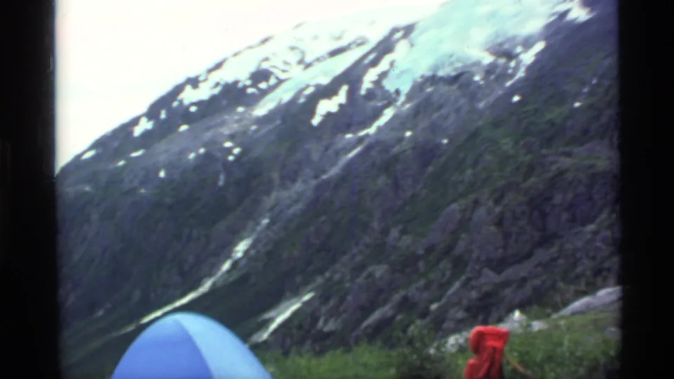 1977 Alaska home-movie frame looking across a rocky mountain valley, with a blue tent edge and red campsite gear in the foreground.