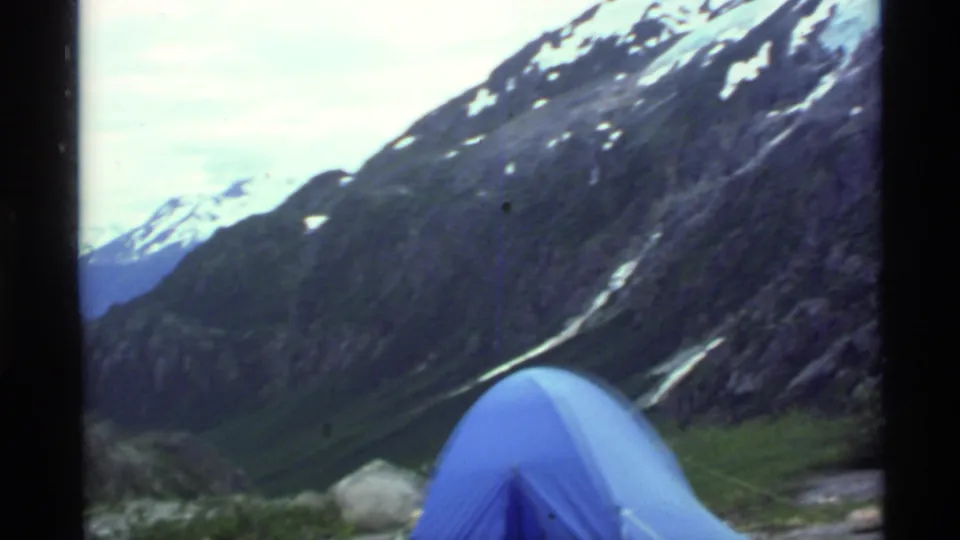 1977 Alaska frame showing a blue dome tent in a small green clearing below rocky, snow-streaked mountain slopes.