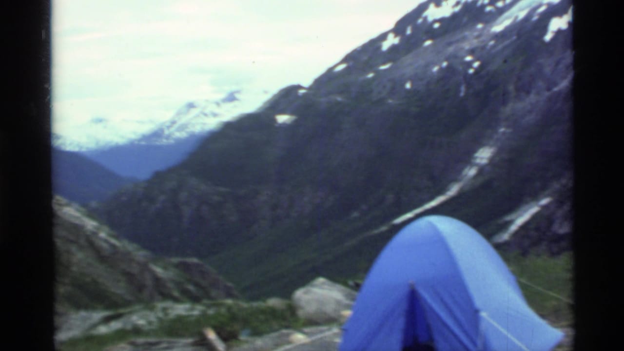 1977 Alaska 8mm frame showing a blue tent at a rocky mountain campsite with a green valley and snow-capped peaks beyond.