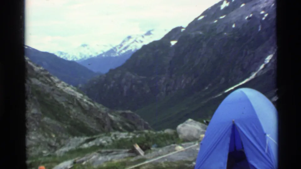 1977 Alaska 8mm frame showing the blue tent, boulders, rocky campsite ground, and a broad valley leading toward snowy peaks.