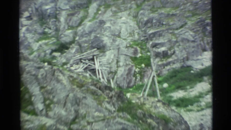 1977 Alaska 8mm frame showing a rough wooden structure or scattered timbers on a rocky, grassy mountain slope.