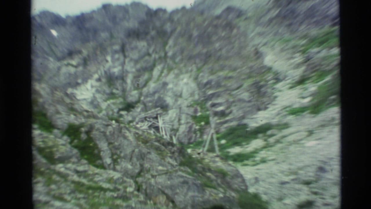 1977 Alaska 8mm frame showing a rugged rocky mountainside with patches of green vegetation under a pale sky.