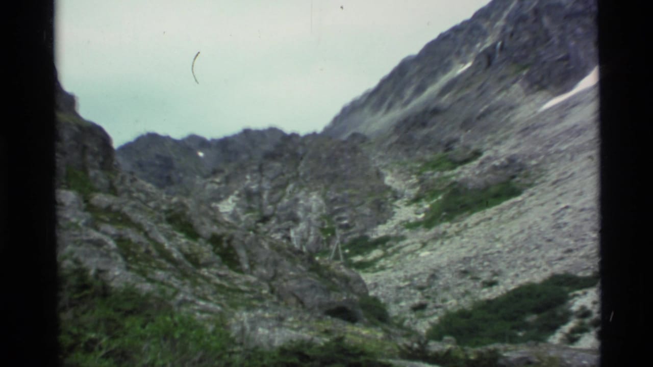 Mountain basin with jagged rock walls, scree, vegetation, and pale sky.