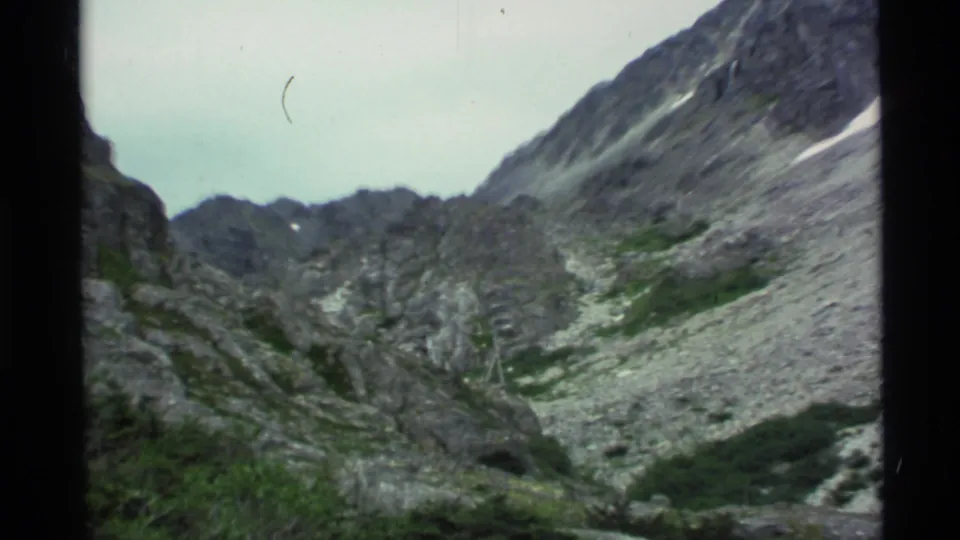 1977 Alaska landscape frame with jagged gray rock walls, scree, low greenery, and an overcast sky.