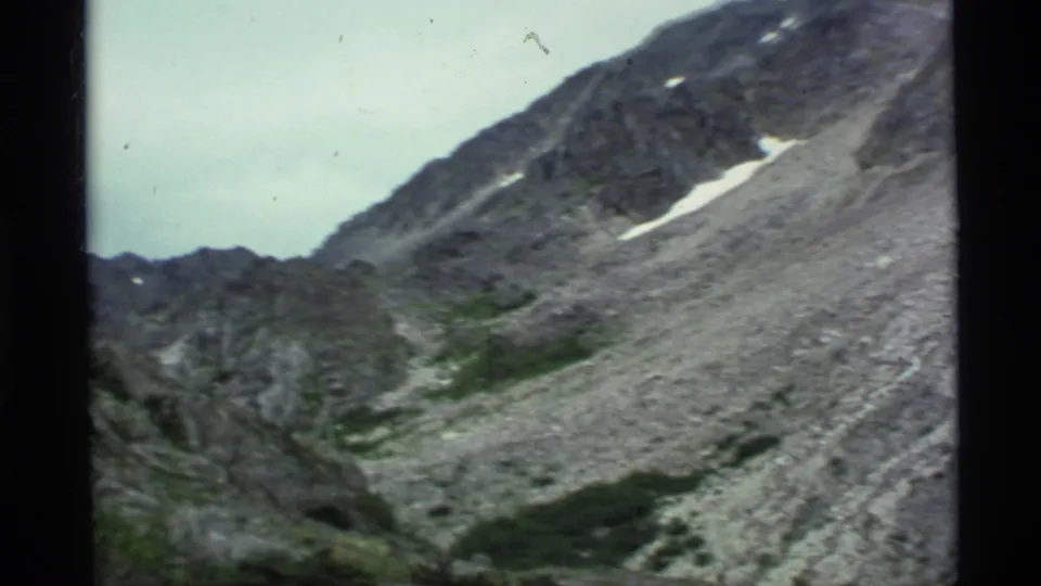1977 Alaska 8mm frame showing gray scree below a steep ridge, with small snow patches and low green vegetation.