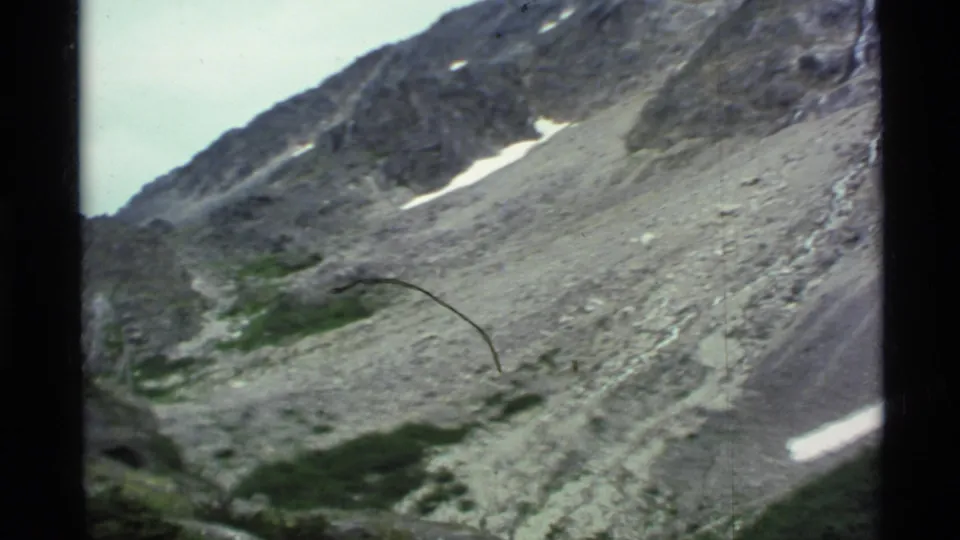 1977 Alaska mountain frame with scree, patches of snow, low greenery, and a visible dark film scratch near the lower center.