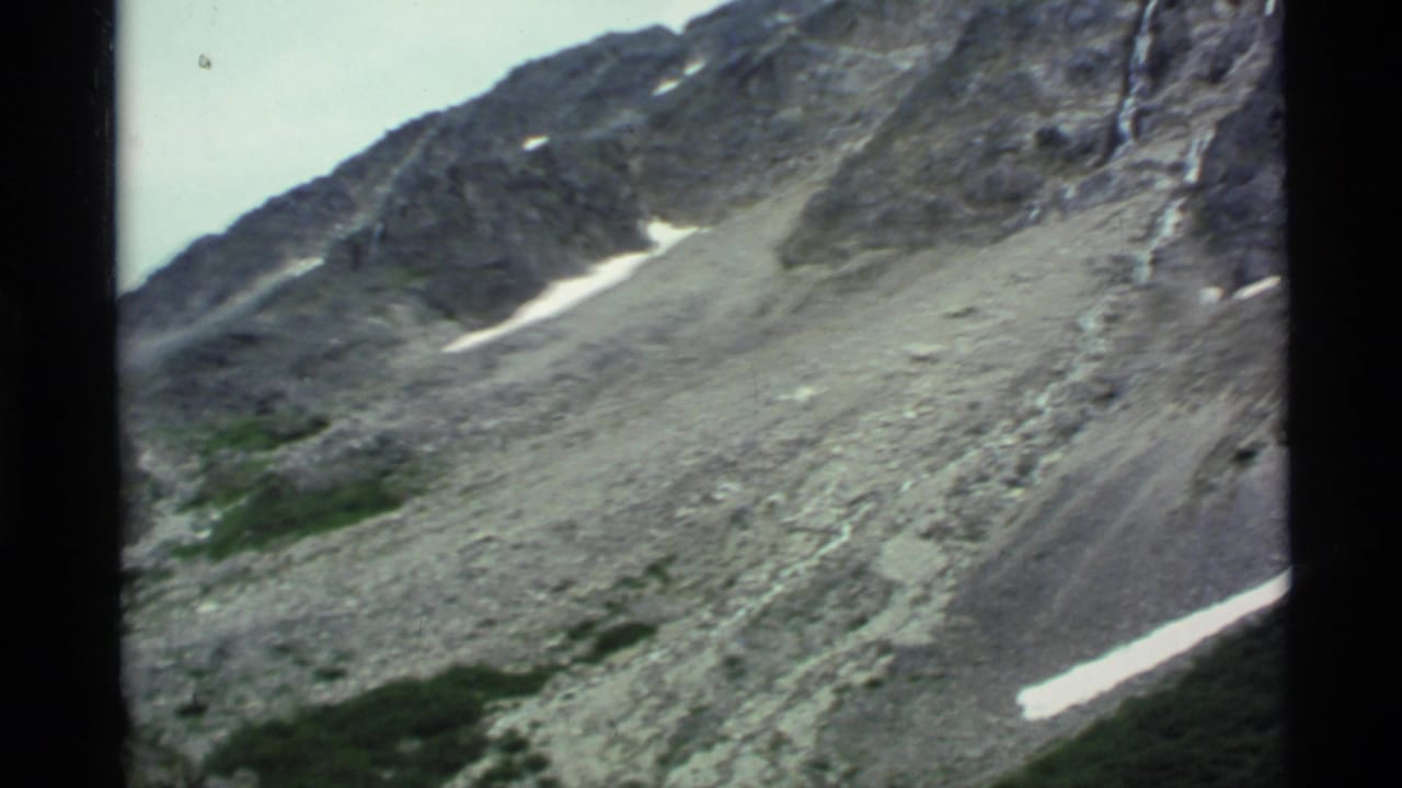 Rocky mountain slope with small snow patches and green vegetation within black 8mm frame borders.