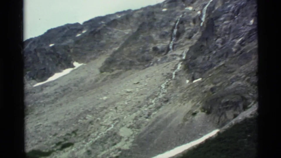 1977 Alaska 8mm frame with a rugged mountain wall, loose scree, small snow fields, and dark projector borders.