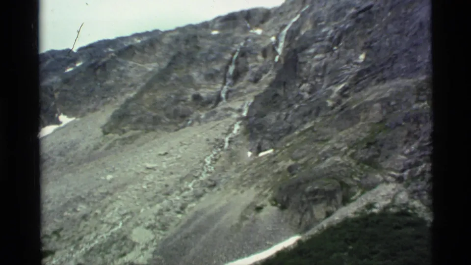 1977 Alaska home-movie frame showing a rocky alpine slope with snow remnants, green brush, and soft film grain.