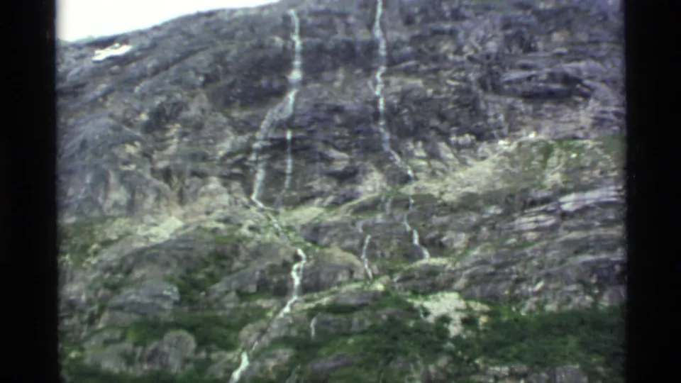 1977 Alaska 8mm frame showing a thin cascade descending a rugged rock face above green alpine vegetation.