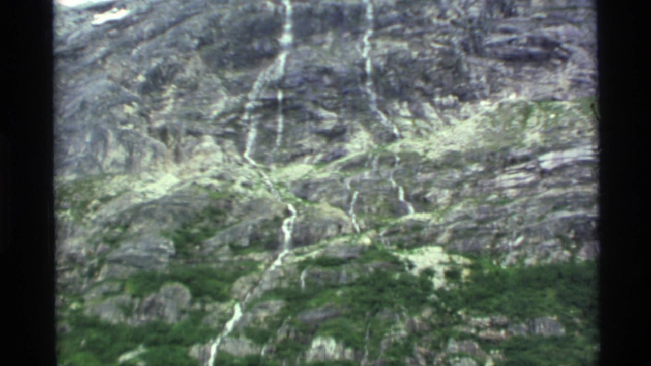 Rocky mountain slope with green vegetation and thin waterfalls visible through film-frame borders.