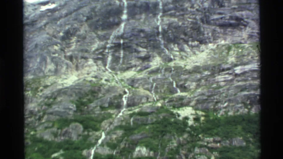 1977 Alaska 8mm frame showing a rugged gray rock face with scattered greenery and narrow white waterfall streaks.