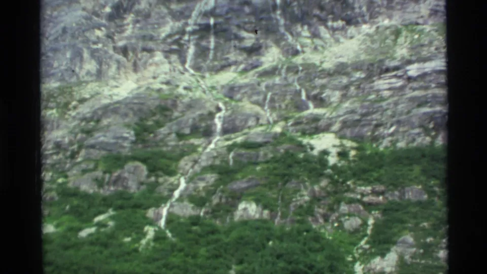 1977 Alaska 8mm view of a steep mountain face with green patches and multiple narrow waterfalls descending the rock.