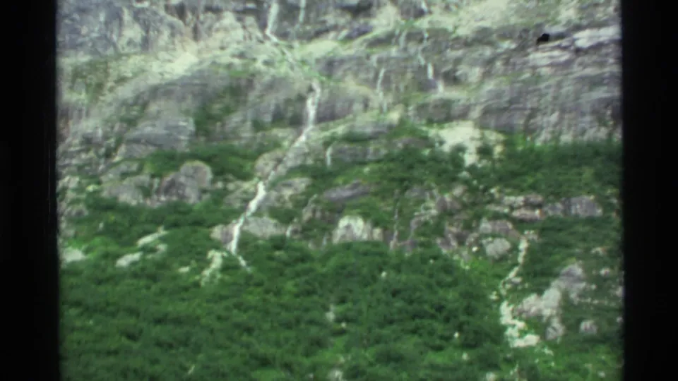 1977 Alaska 8mm frame with motion-softened view of green vegetation below a rocky slope marked by narrow watercourses.