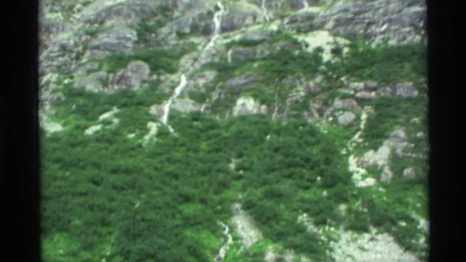 1977 Alaska 8mm frame showing green mountainside vegetation, exposed rock, pale runoff paths, and a narrow waterfall.
