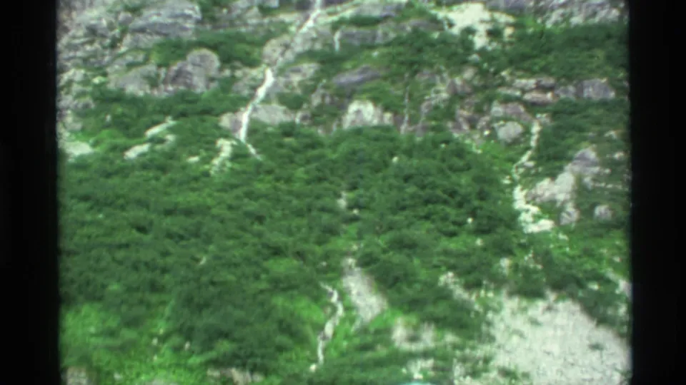 1977 Alaska home-movie frame showing a green rocky mountainside with a thin waterfall or stream descending through pale stone.
