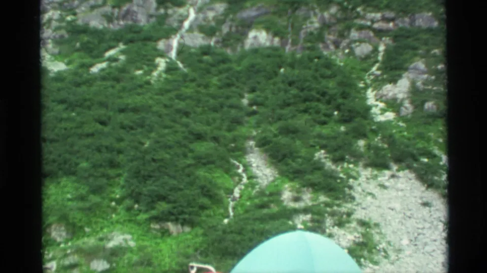 1977 Alaska camping footage frame with a rocky green mountainside and the edge of a pale turquoise tent entering the foreground.