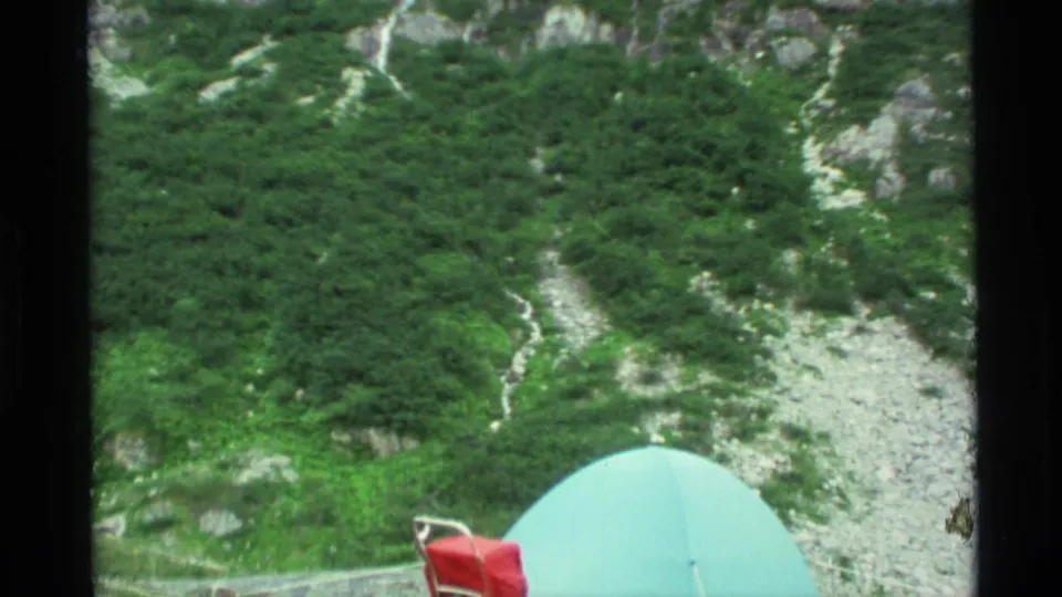 1977 Alaska home-movie frame showing a pale turquoise tent and red camping fabric below a steep, brushy mountain slope.