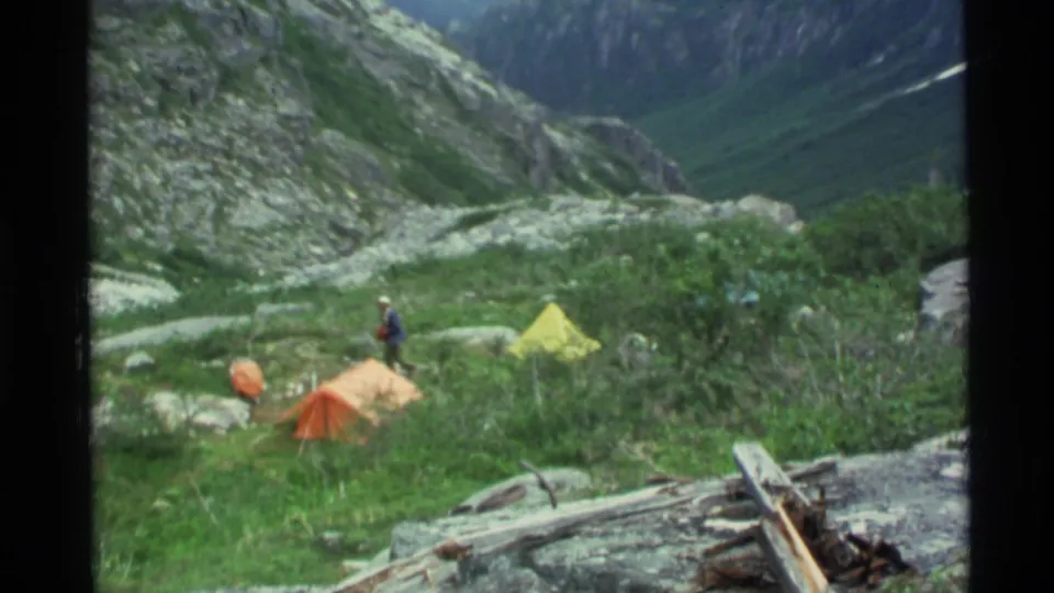 1977 Alaska camping frame showing orange and yellow tents on a grassy slope, with a distant person standing among rocks and shrubs.
