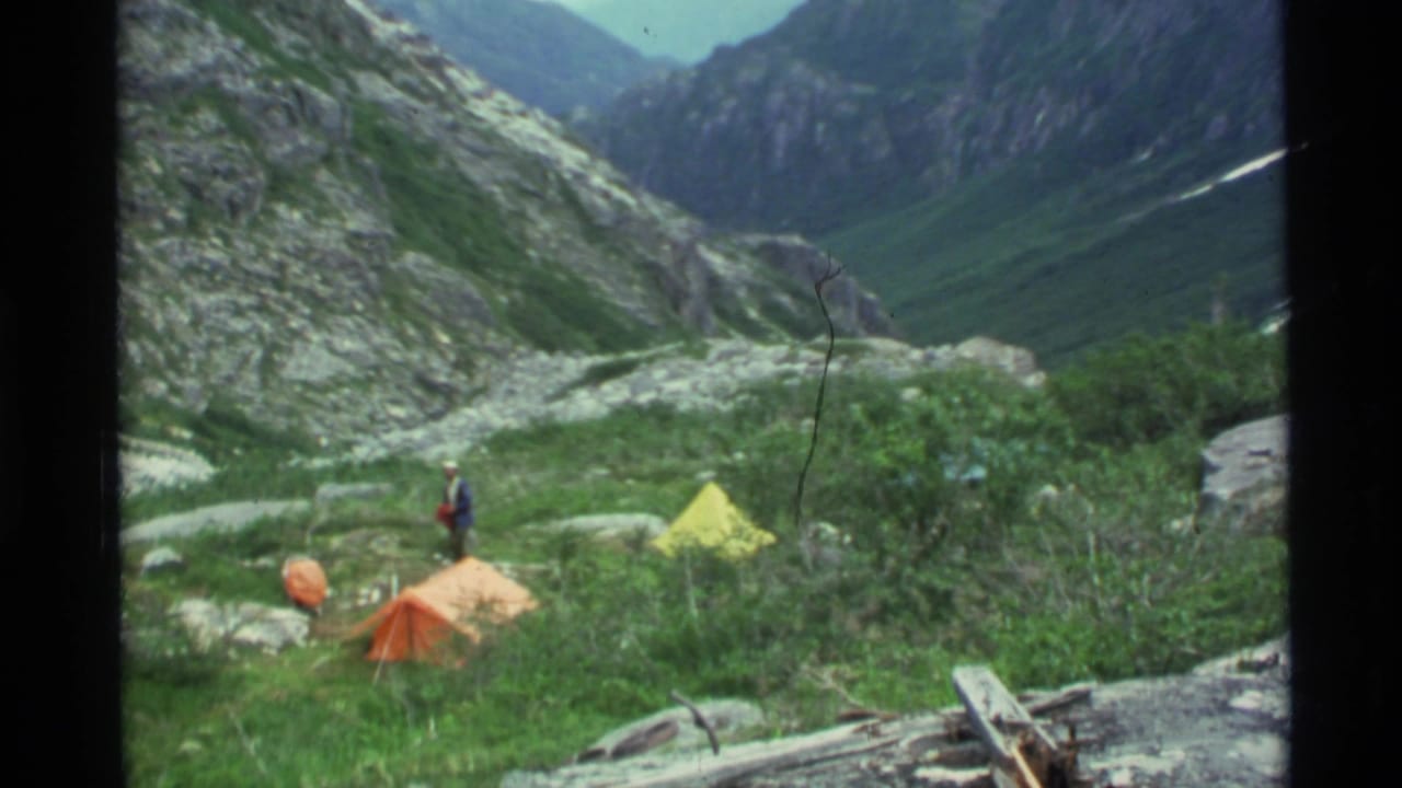 1977 Alaska 8mm frame showing tents and a small camper in a rocky mountain valley with distant snow-capped peaks.