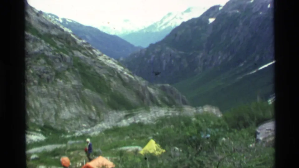 1977 Alaska home-movie view of a camper near bright tents beneath steep rocky mountains and snow in the distance.