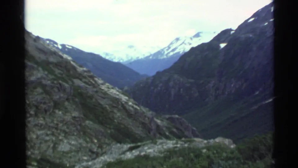 1977 Alaska 8mm frame looking across rocky alpine terrain toward distant snow-capped mountains.
