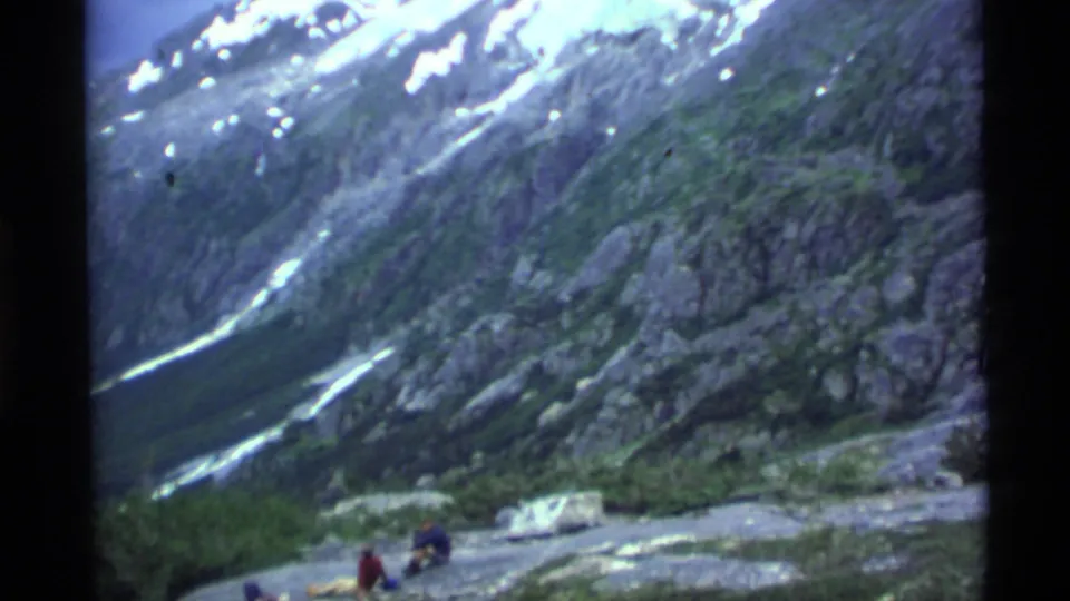 1977 Alaska 8mm frame showing a steep rocky mountainside with snow streaks and distant seated people on open ground.