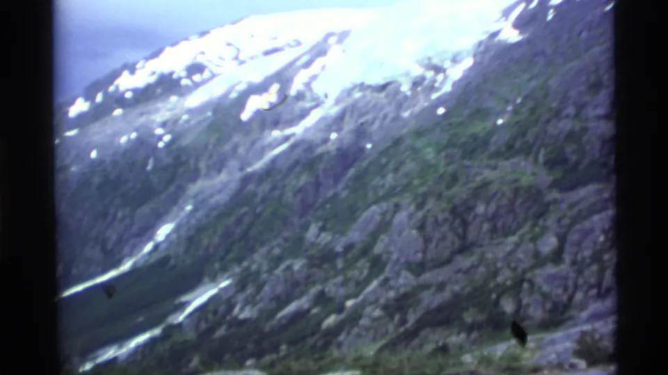 1977 Alaska 8mm view of a rugged mountainside with diagonal snow or glacier streaks and muted green vegetation.