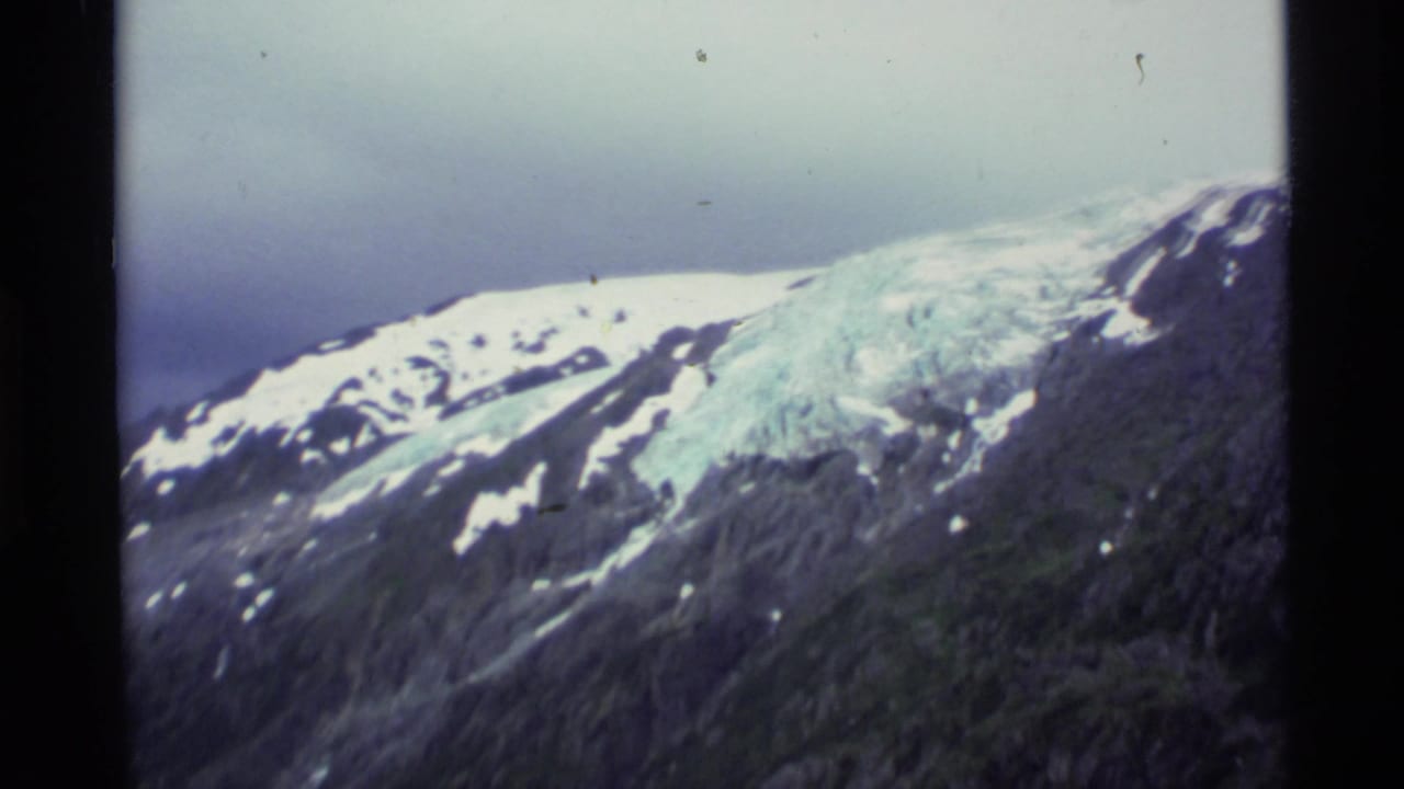 1977 Alaska 8mm frame showing a snow-capped ridge, blue-white glacier, and dark mountain slope beneath a muted sky.