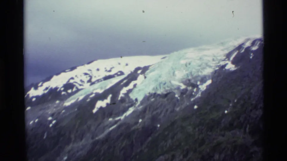 1977 Alaska 8mm frame showing a snow-capped ridge, blue-white glacier, and dark mountain slope beneath a muted sky.