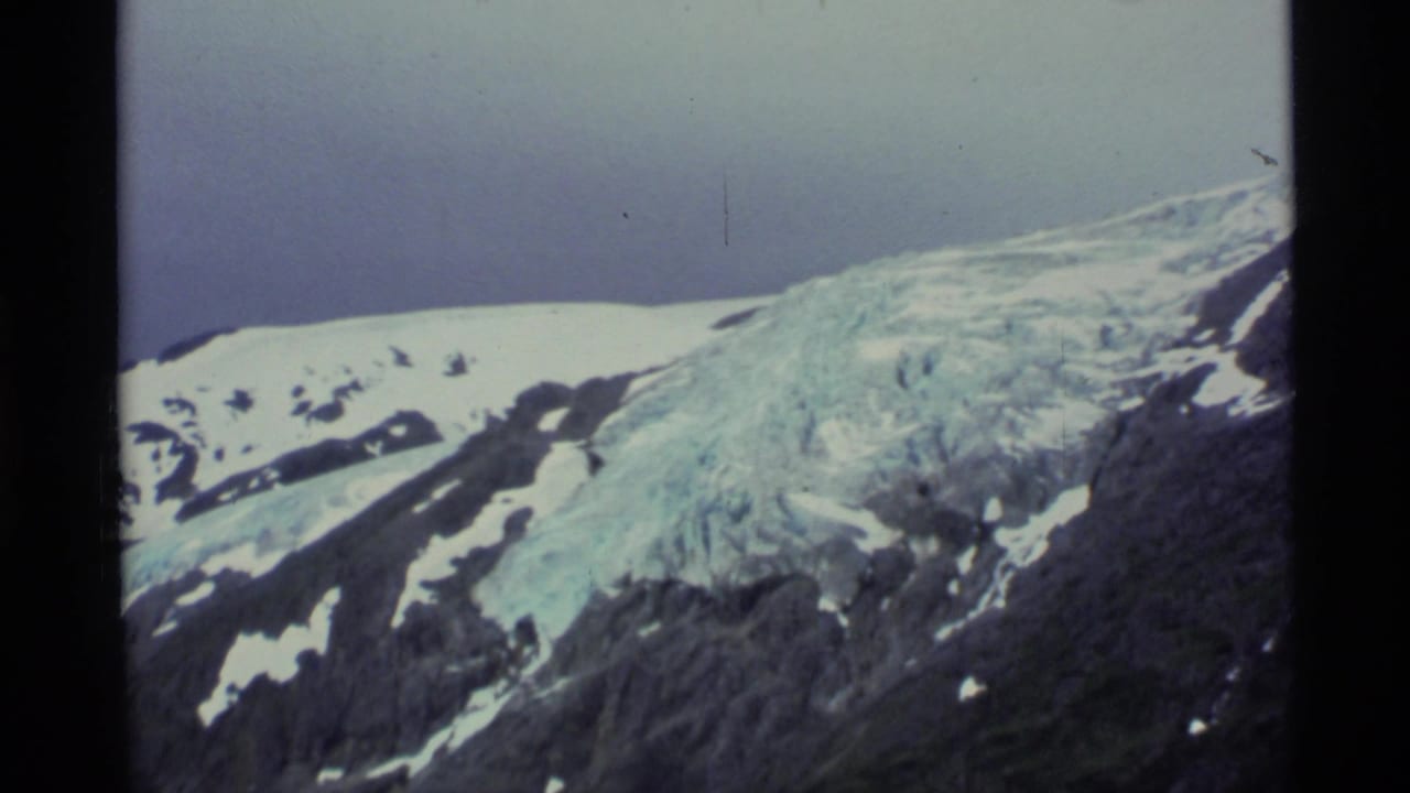 1977 Alaska home-movie frame with glacier ice, snow patches, dark rock, and a pale sky in handheld 8mm color.