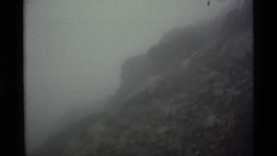 1977 Alaska 8mm frame where thick white fog obscures much of the mountain terrain and leaves only a dark slope partly visible.