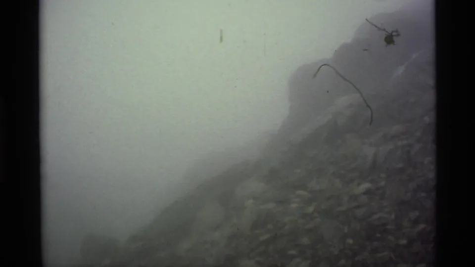 1977 Alaska 8mm frame showing dense fog overtaking a rocky mountain slope, with only faint terrain and film marks remaining visible.