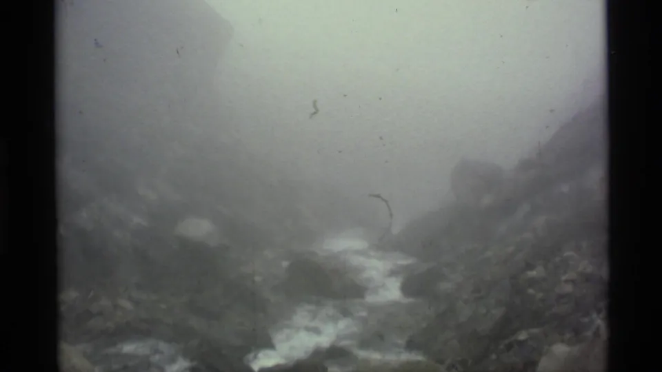Fog hangs over an Alaskan mountain valley as a pale stream cuts through rocks in this 1977 8mm frame.