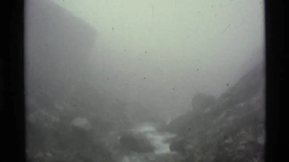 A hazy 1977 8mm view of Alaska mountain terrain shows a stream and boulders fading into thick fog.