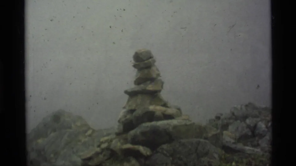 A small stone cairn stands on rocky Alaskan terrain under heavy fog in this 1977 8mm home-movie frame.