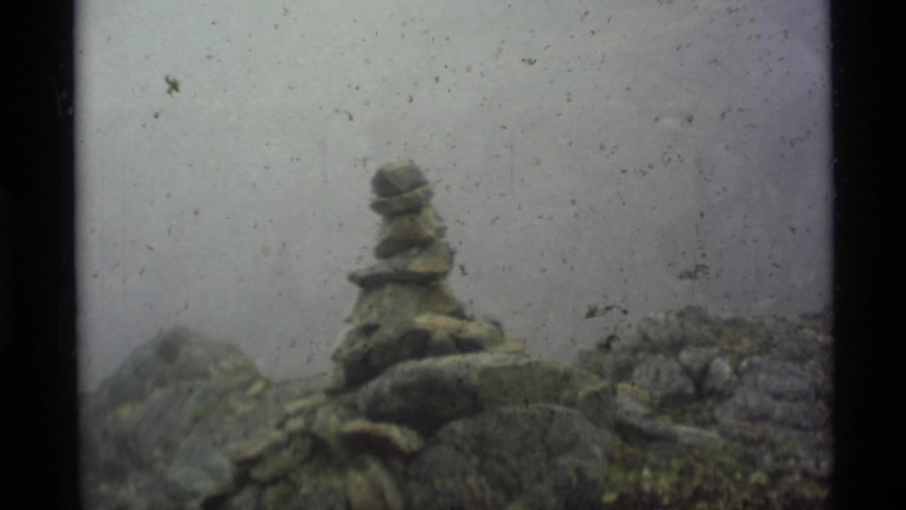 1977 Alaska 8mm frame showing a small stacked stone cairn amid wet rocks and mist, with visible dust and soft focus.